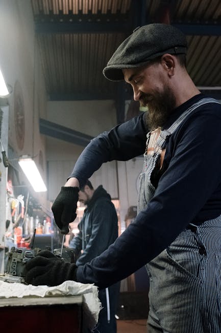 Auto mechanic repairing a vehicle part in a well-lit garage workshop, wearing gloves and cap.
