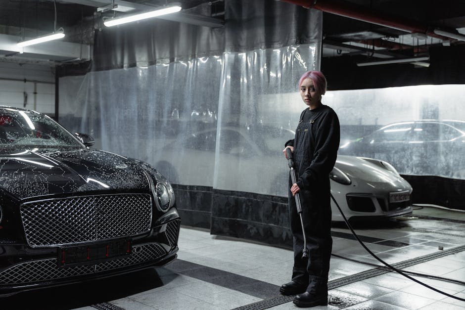 A woman with pink hair operates a power spray in a carwash garage with luxury cars.
